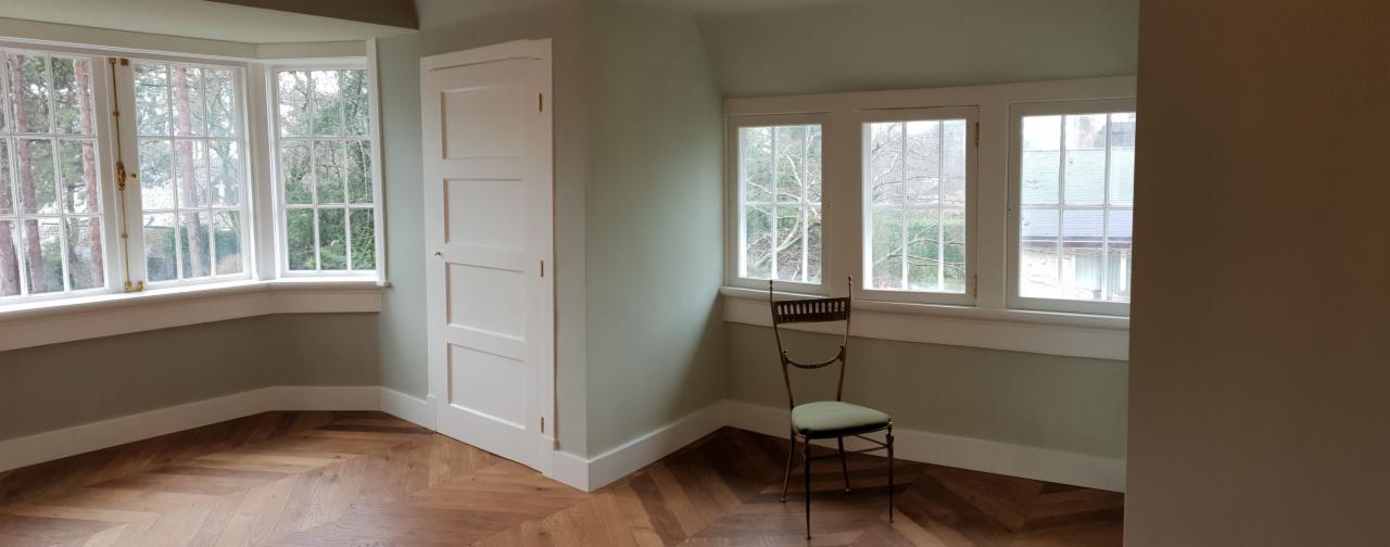 Jasne wnętrze po odświeżeniu z parkietem ułożonym w jodełkę, świeżo pomalowanymi ścianami w odcieniach zieleni i białymi listwami przypodłogowymi, z krzesłem przy oknie.