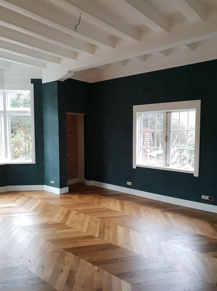 Pusty pokój po remoncie z ciemnozielonymi ścianami, białymi oknami i parkietem ułożonym we wzór jodełki francuskiej, widoczne gniazdka elektryczne bez osłon.