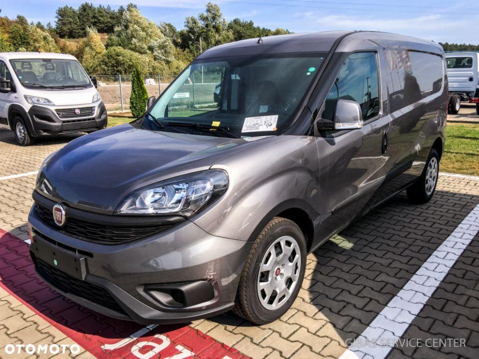 Szary samochód dostawczy Fiat Doblo Cargo na parkingu z kostki brukowej, widoczny z przodu pod kątem, z innym białym busem w tle i drzewami w oddali.