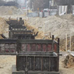 Szeroki kadr budowy fundamentów z użyciem metalowych szalunków w wykopie, widoczna koparka i składowany urobek ziemi.