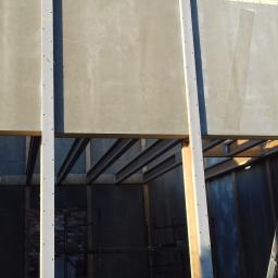 F.U.H Rene-Bud - Montaż prefabrykowanych ścian zewnętrznych na budowie domu; widoczne stalowe belki stropowe, drabina oparta o ścianę i fragment rusztowania wewnątrz budynku.