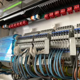 Szczegółowe ujęcie rozdzielnicy elektrycznej z licznymi przewodami oznaczonymi etykietami, listwami zaciskowymi w kolorze szarym i niebieskim oraz elementami zabezpieczającymi.