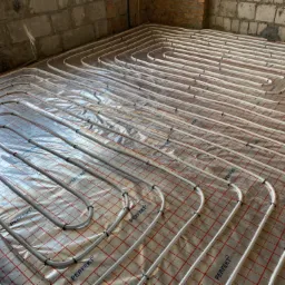 Białe rury instalacji grzewczej podłogowej ułożone w pętle na folii z czerwoną kratką, przymocowane czarnymi klipsami, widok z góry w surowym stanie budynku.