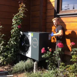 Instalator w niebieskich rękawicach daje kciuk w górę obok zainstalowanej pompy ciepła na tle drewnianego domu z kwitnącymi różami.