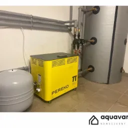 Żółty piec Pereko 10kW obok szarego zbiornika wyrównawczego i ocieplonego zasobnika CWU z podłączeniami hydraulicznymi, rury w izolacji na ścianie, brudna podłoga z płytek.