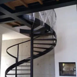 Metalowa, spiralna klatka schodowa w surowym wnętrzu, zabezpieczona folią na górze, z betonową ścianą w tle i okrągłą drewnianą podstawą.