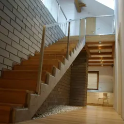 Wewnętrzne schody drewniane z betonowymi stopniami i balustradą ze stali nierdzewnej, ściana z cegły, pod schodami wysypane kamienie, drewniana podłoga, w tle widoczne pomieszczenie z oknem...