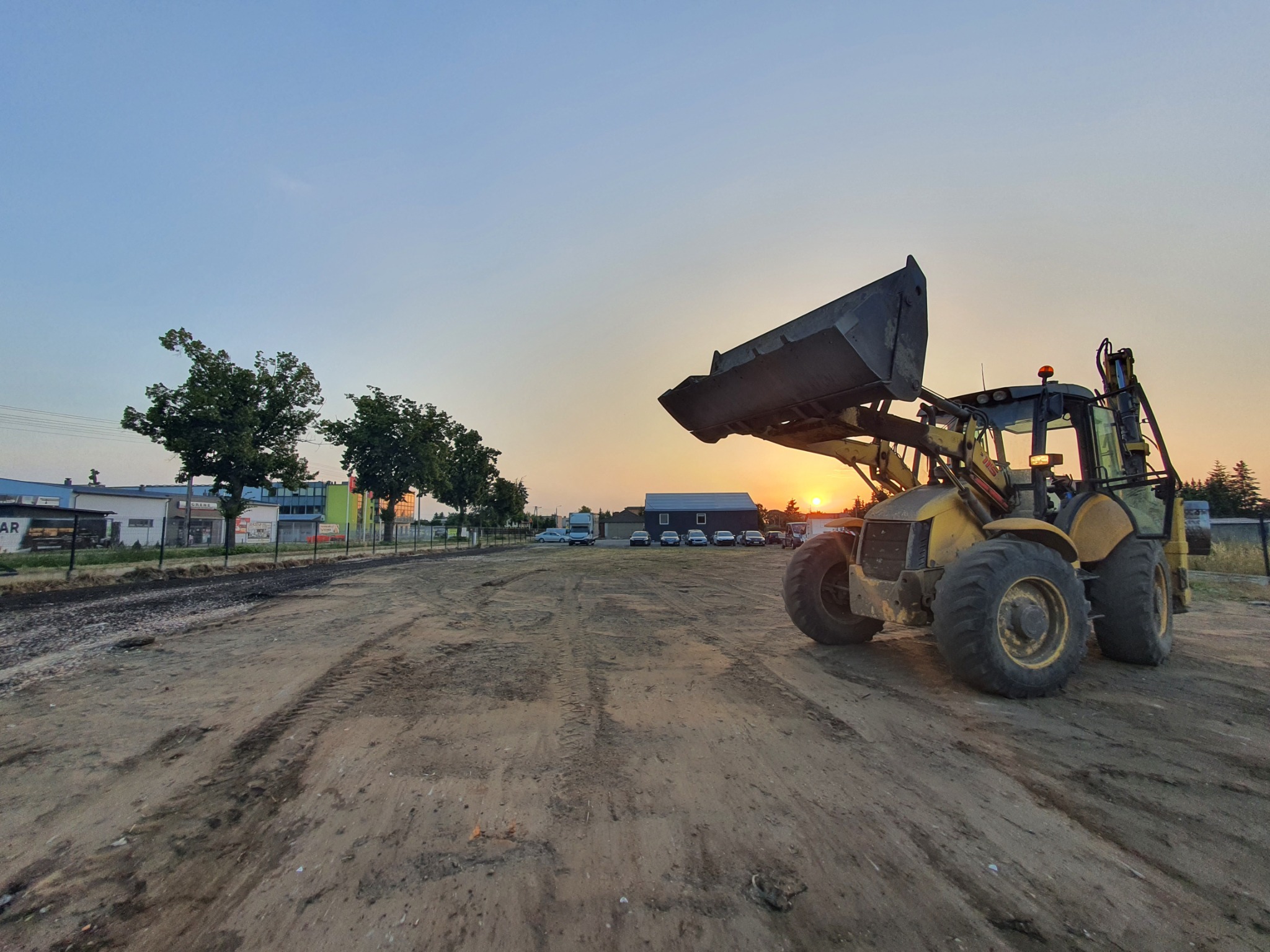 Żółta koparko-ładowarka z podniesioną łyżką stoi na placu budowy o zmierzchu; w tle budynki i zaparkowane samochody.