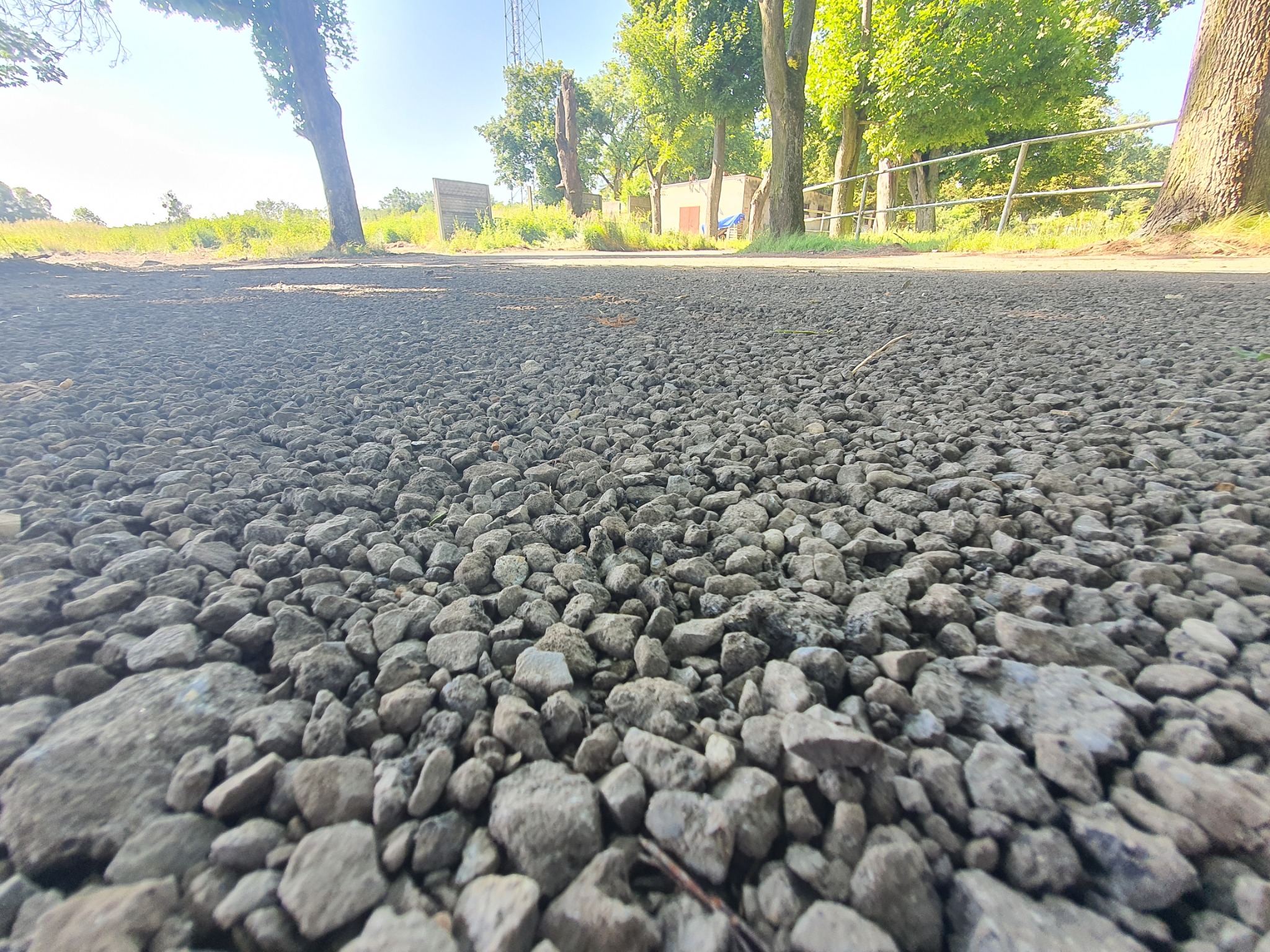 Nisko położone ujęcie szarego, drobnego kruszywa na utwardzonej drodze gruntowej z widocznymi drzewami i zielenią w tle.