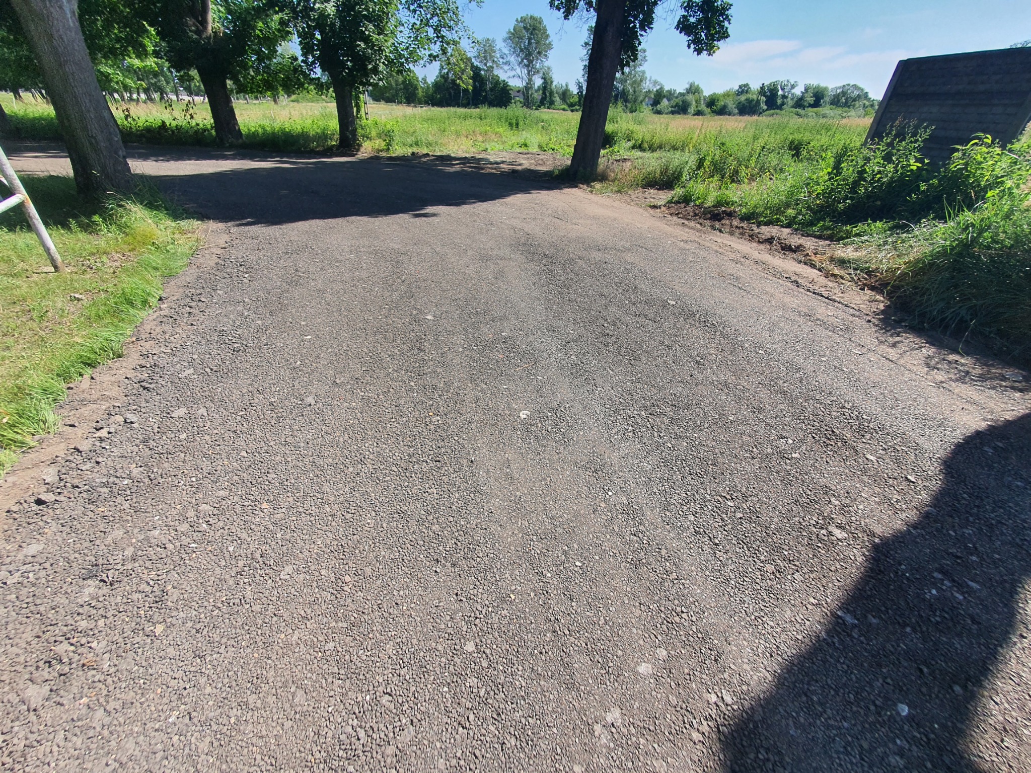 Świeżo utwardzona droga gruntowa z drobnym kruszywem, widok z poziomu gruntu, z drzewami po bokach i drabiną w tle.