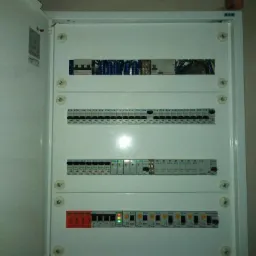 Otwarta, biała skrzynka rozdzielcza z widocznymi bezpiecznikami i przewodami. Górna część skrzynki zawiera listwę z niebieskimi zaciskami. Niektóre włączniki są w pozycji ON, sygnalizowane...