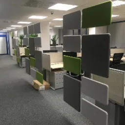 Nowoczesne biuro z panelami akustycznymi w odcieniach szarości i zieleni, wiszącymi pionowo pomiędzy stanowiskami pracy, widok na puste biurka i krzesła biurowe, w tle drzwi i rośliny doniczkowe.