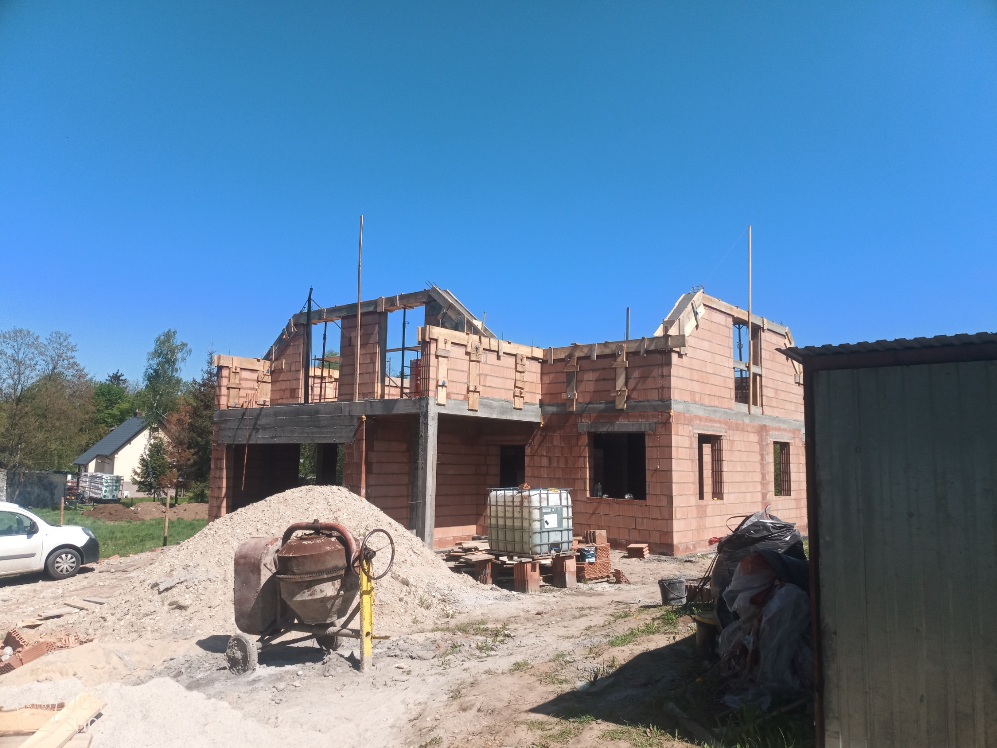 Budowa domu w stanie surowym otwartym: widoczne ściany z czerwonej cegły, częściowo wykonana więźba dachowa, stos piasku, betoniarka i zbiornik na wodę na placu budowy w słoneczny dzień.