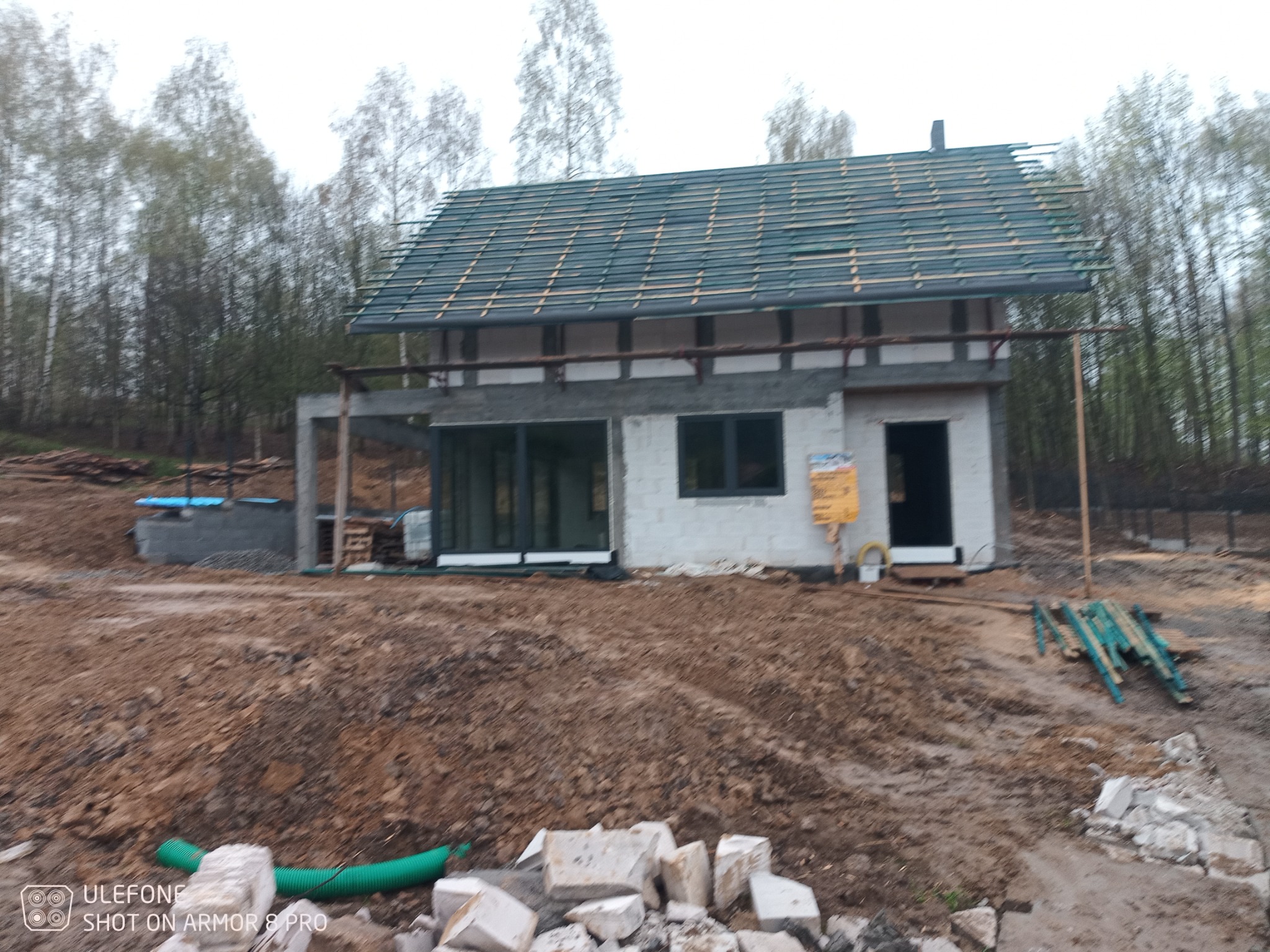 Niedokończony dom jednorodzinny z częściowo położonym dachem z dachówki ceramicznej, widoczne rusztowania i przygotowany teren wokół budynku.