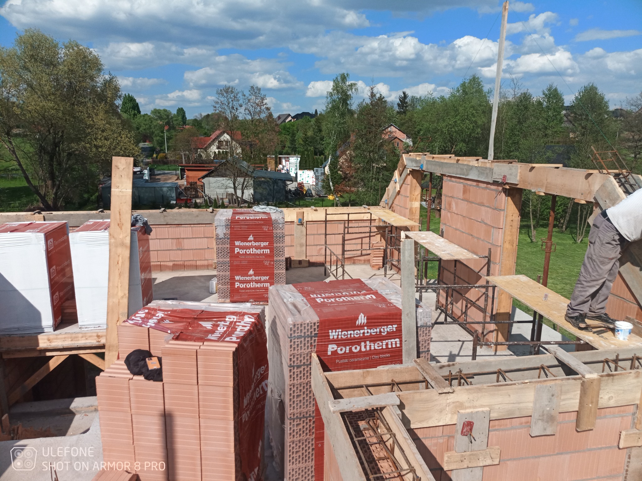 Budowa domu w trakcie wznoszenia ścian z czerwonej cegły Porotherm, widoczne rusztowania, drewniane szalunki i materiały budowlane na placu budowy w słoneczny dzień.