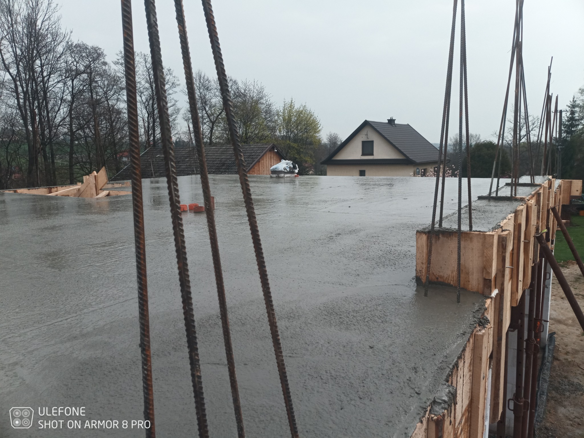 Zalany betonem fundament płyty, z widocznym zbrojeniem i szalunkiem drewnianym, w tle zabudowania gospodarcze i dom jednorodzinny w pochmurny dzień.