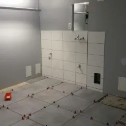 Łazienka w trakcie remontu: szare płytki podłogowe układane z użyciem krzyżyków dystansowych, białe płytki ścienne częściowo położone, widoczne instalacje wodno-kanalizacyjne.