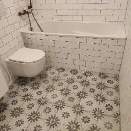 Nowoczesna łazienka z białą armaturą, wanną obudowaną białymi płytkami, wiszącą toaletą z bidetką, i podłogą w geometryczne, kwiatowe wzory.