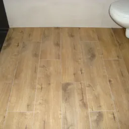 Ułożone panele podłogowe imitujące drewno w łazience, widoczny fragment toalety i czarna ściana z płytek.