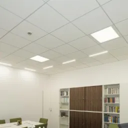 Sufit podwieszany z płyt kartonowo-gipsowych z wbudowanym oświetleniem LED w biurze lub sali konferencyjnej, widoczny czujnik dymu, białe ściany i regały z książkami.