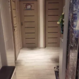 Wnętrze korytarza z trzema drewnianymi drzwiami z poziomymi panelami, z których jedne ozdobione naklejką z motywem dziecięcym, podłoga wyłożona jasnymi panelami imitującymi drewno, widoczny...