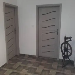 Dwa szare skrzydła drzwiowe z poziomymi, czarnymi zdobieniami, ustawione w białym pomieszczeniu z wzorzystą podłogą, obok stoi kołowrotek.