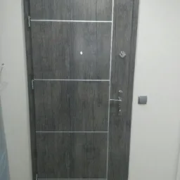 Szare drzwi wejściowe z okleiną imitującą drewno, z trzema poziomymi panelami oddzielonymi srebrnymi liniami, widoczny zamek, wizjer i klamka.