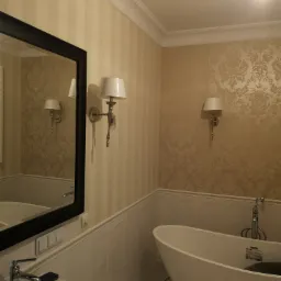 Elegancka łazienka z wolnostojącą wanną, wzorzystą tapetą i kinkietami w stylu retro, odbicie w lustrze podkreśla symetrię pomieszczenia.