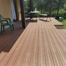 Nowoczesny taras z desek kompozytowych w odcieniach brązu, z widocznym zadaszeniem, meblami ogrodowymi i zielenią w tle, częściowo zacieniony.