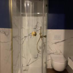 Nonamee - Nowoczesna kabina prysznicowa z elementami w kolorze złotym, marmurowe płytki na ścianach, wisząca toaleta i drewnopodobna podłoga w łazience.