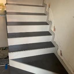 Nonamee - Schody wykończone płytkami w odcieniach szarości i bieli, widoczne ślady po remoncie na ścianie, częściowo położona podłoga i narzędzia w tle.