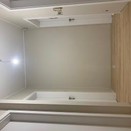 Nonamee - Wnętrze w trakcie prac: ściany po szpachlowaniu i malowaniu na jasny beż, widoczne fragmenty podłogi z paneli imitujących drewno, surowa instalacja oświetleniowa z żarówką bez oprawy, oraz białe...