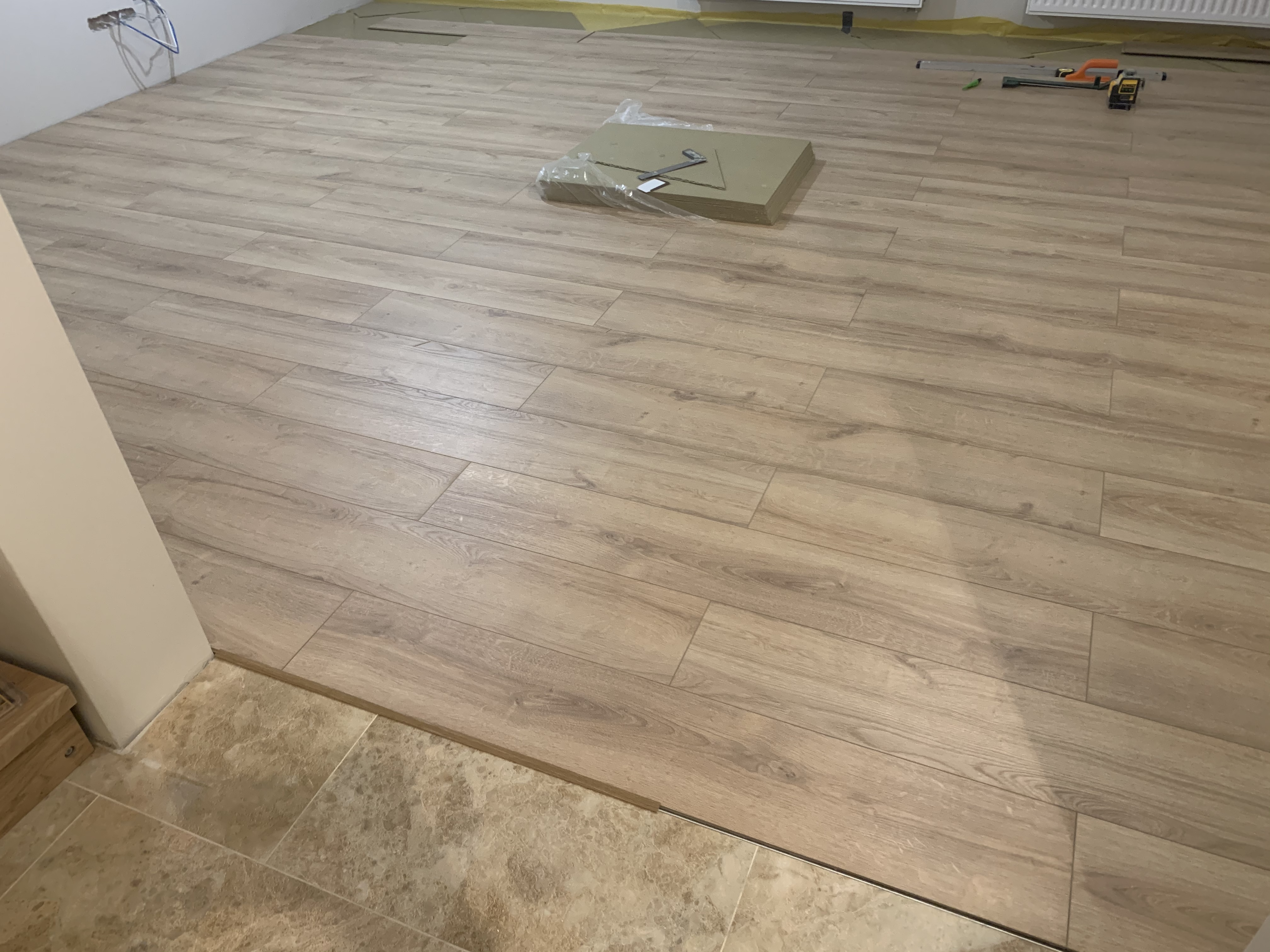 Układanie paneli podłogowych w jasnym odcieniu drewna, widoczne narzędzia i materiały przygotowane do montażu, przejście do płytek podłogowych przy ścianie.