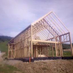 Surowy szkielet drewnianego domu w trakcie budowy, widoczny na tle pag&oacute;rkowatego terenu i błękitnego nieba, z logo firmy na zaparkowanym obok samochodzie dostawczym.