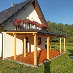 Jasny dom z ciemnym dachem i drewnianą werandą z balustradą, balkon z czerwonymi kwiatami, widok na zielony ogród i las.