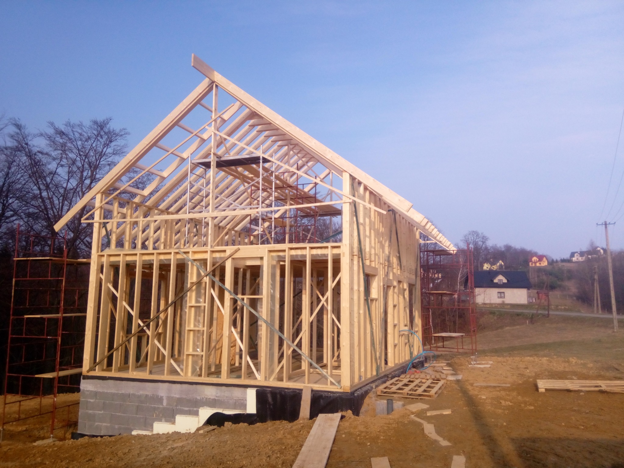 Wznoszenie konstrukcji domu w technologii szkieletowej na fundamencie z bloczków betonowych, widoczne rusztowania i elementy konstrukcyjne dachu, w tle zabudowa jednorodzinna na wzgórzu.