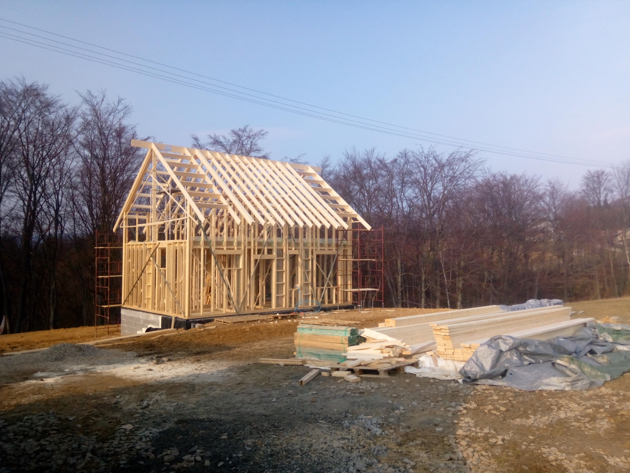 Konstrukcja szkieletowa domu w trakcie budowy, widoczne rusztowania i stos drewna na placu budowy, drzewa w tle