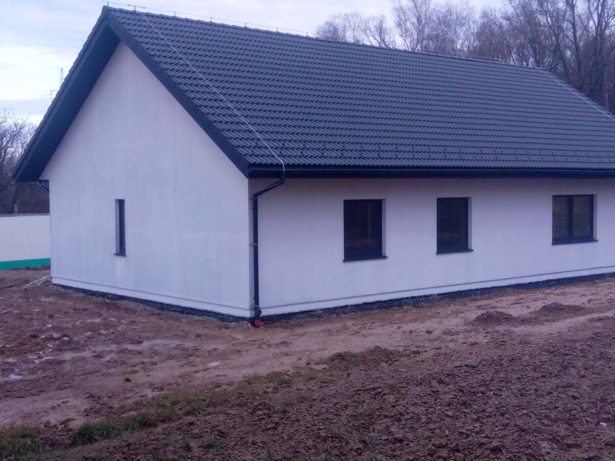 Nowo wybudowany dom szkieletowy z ciemną dachówką i białymi ścianami zewnętrznymi, widok z zewnątrz na tle drzew bez liści i błotnistego terenu.