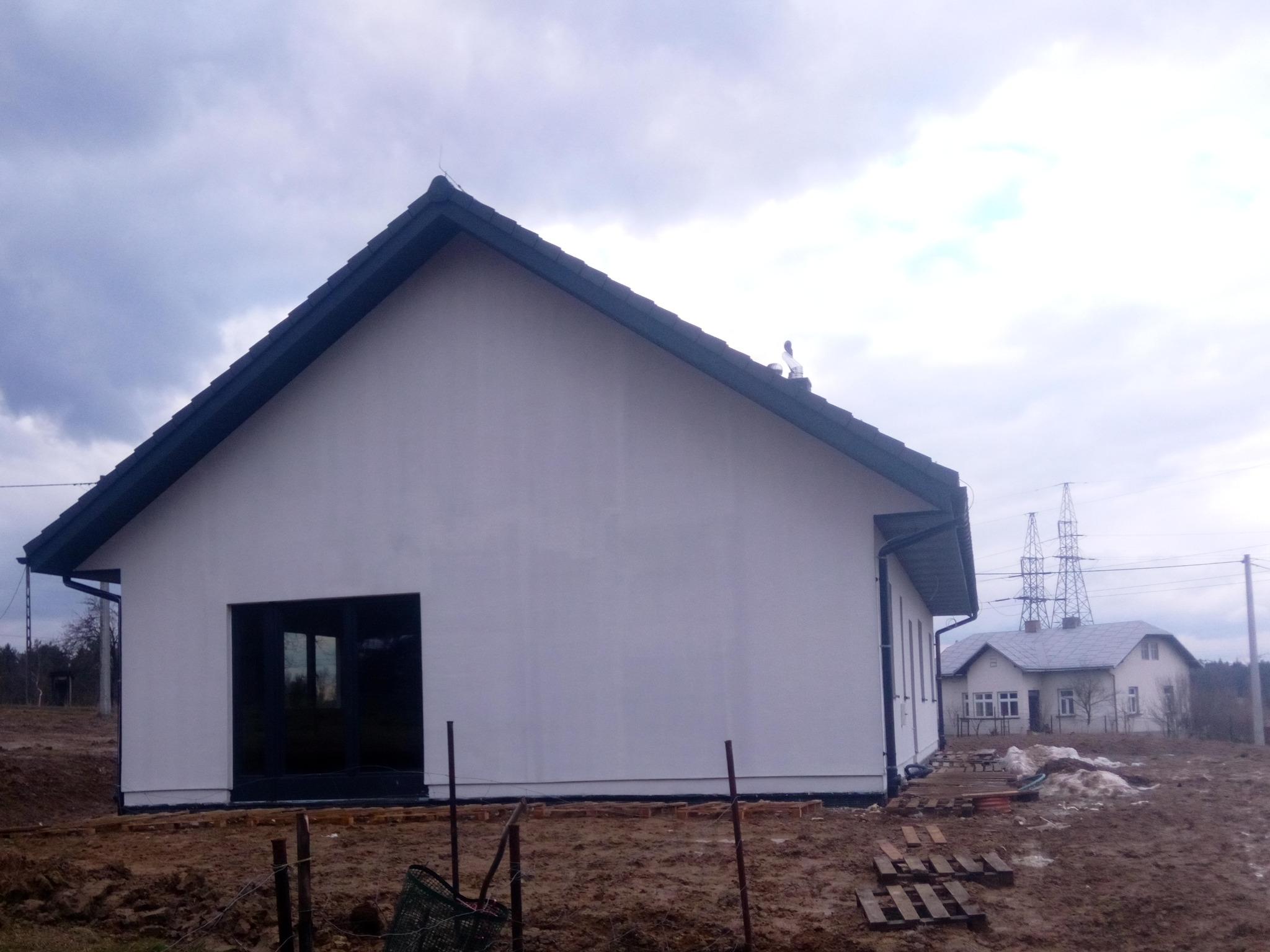 Surowy stan nowo wybudowanego, otynkowanego domu z ciemnymi oknami i dachem, widok z boku na tle pochmurnego nieba i innego domu w oddali.