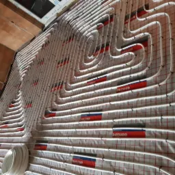 Białe rury ogrzewania podłogowego ułożone spiralnie na izolacji z czerwono-białą siatką, widoczne elementy mocujące i fragment ściany z cegły.