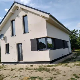 Nowoczesny dom jednorodzinny z beżową elewacją i czarnymi oknami, widok z boku na świeżo wykończoną fasadę z satelitą na ścianie, teren wokół domu jeszcze niezagospodarowany.