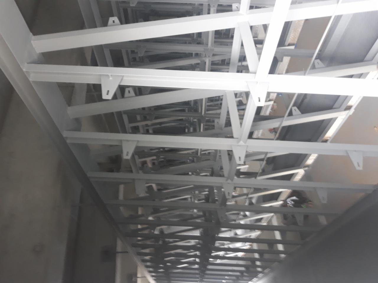 Biała konstrukcja stalowa pod sufitem, widok z dołu, elementy połączone spawami i śrubami, równomiernie pokryte farbą.