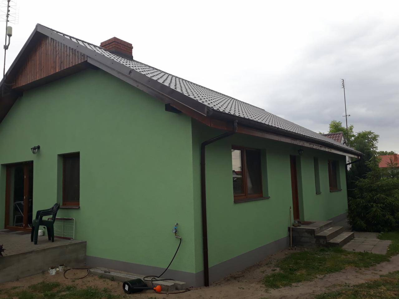 Świeżo pomalowana elewacja domu jednorodzinnego w odcieniach zieleni, z widocznymi śladami prac wykończeniowych, w tle pochmurne niebo.