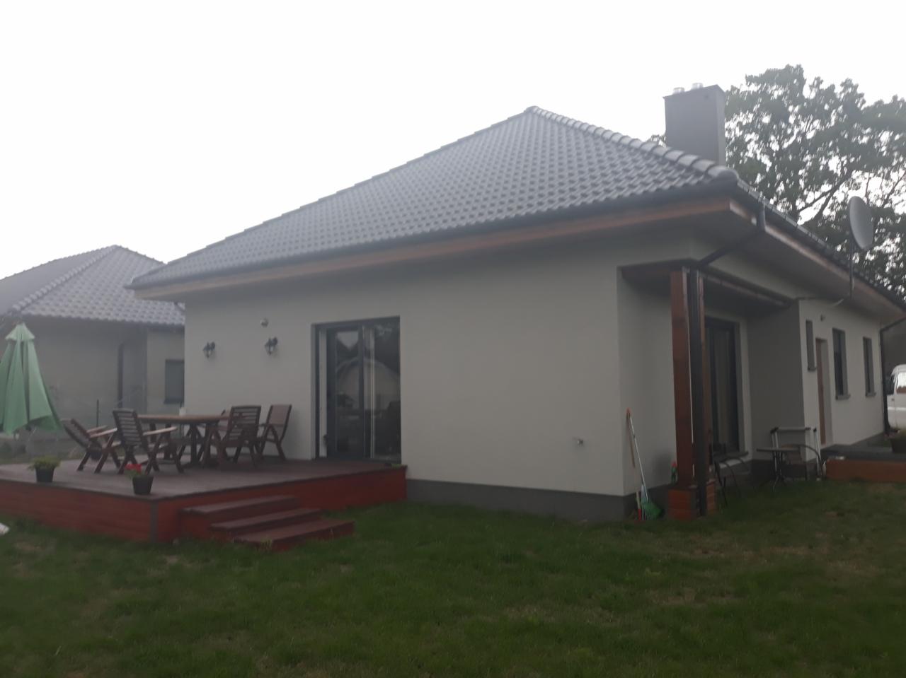 Elewacja domu jednorodzinnego w jasnym odcieniu szarości, z ciemną dachówką i drewnianym tarasem. Widoczne elementy ogrodu i sąsiedni budynek.