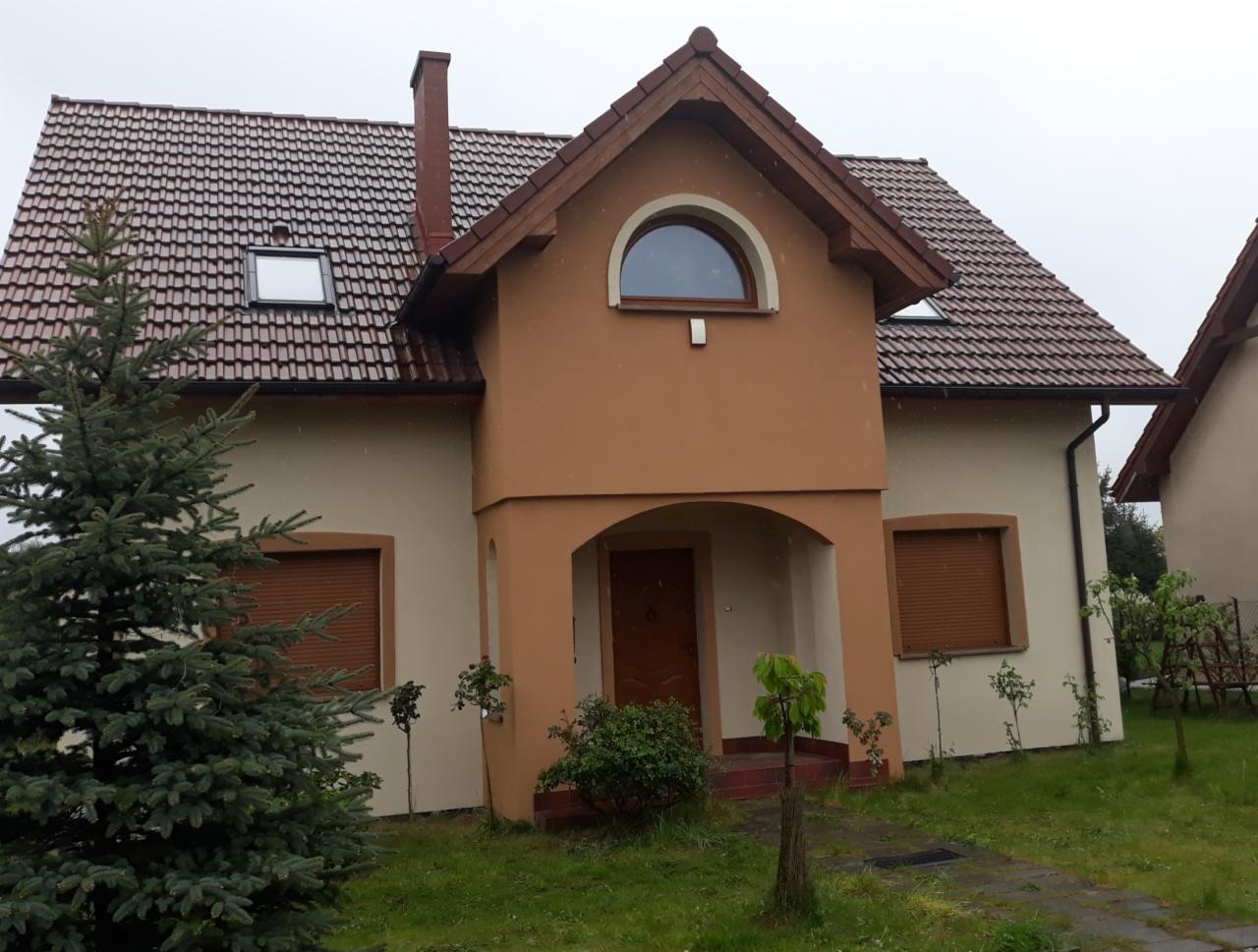 Dwukondygnacyjny dom jednorodzinny z brązową elewacją i dachem z czerwonej dachówki, widoczny w pochmurny dzień z przodu, z zielonym drzewem iglastym po lewej stronie.