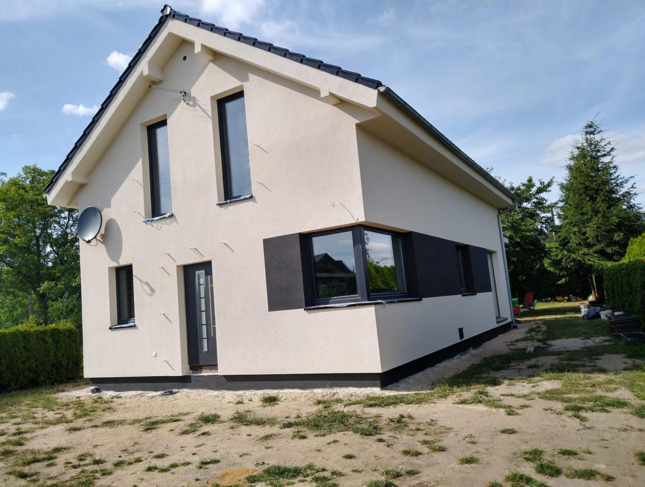 Nowoczesny dom jednorodzinny z beżową elewacją i czarnymi oknami, widok z boku na świeżo wykończoną fasadę z satelitą na ścianie, teren wokół domu jeszcze niezagospodarowany.