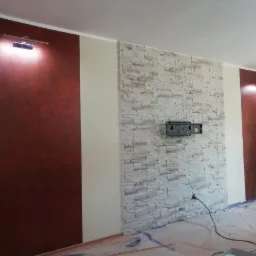 Pokój po remoncie: ściany w odcieniach czerwieni i bieli, fragment ściany wyłożony kamieniem dekoracyjnym, widoczny uchwyt na telewizor i drabina w rogu.