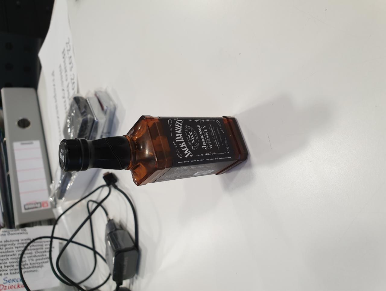 Miniaturowa butelka Jack Daniel's, prawdopodobnie element zestawu upominkowego, leży na białym biurku obok dokumentów, kabli i segregatora.