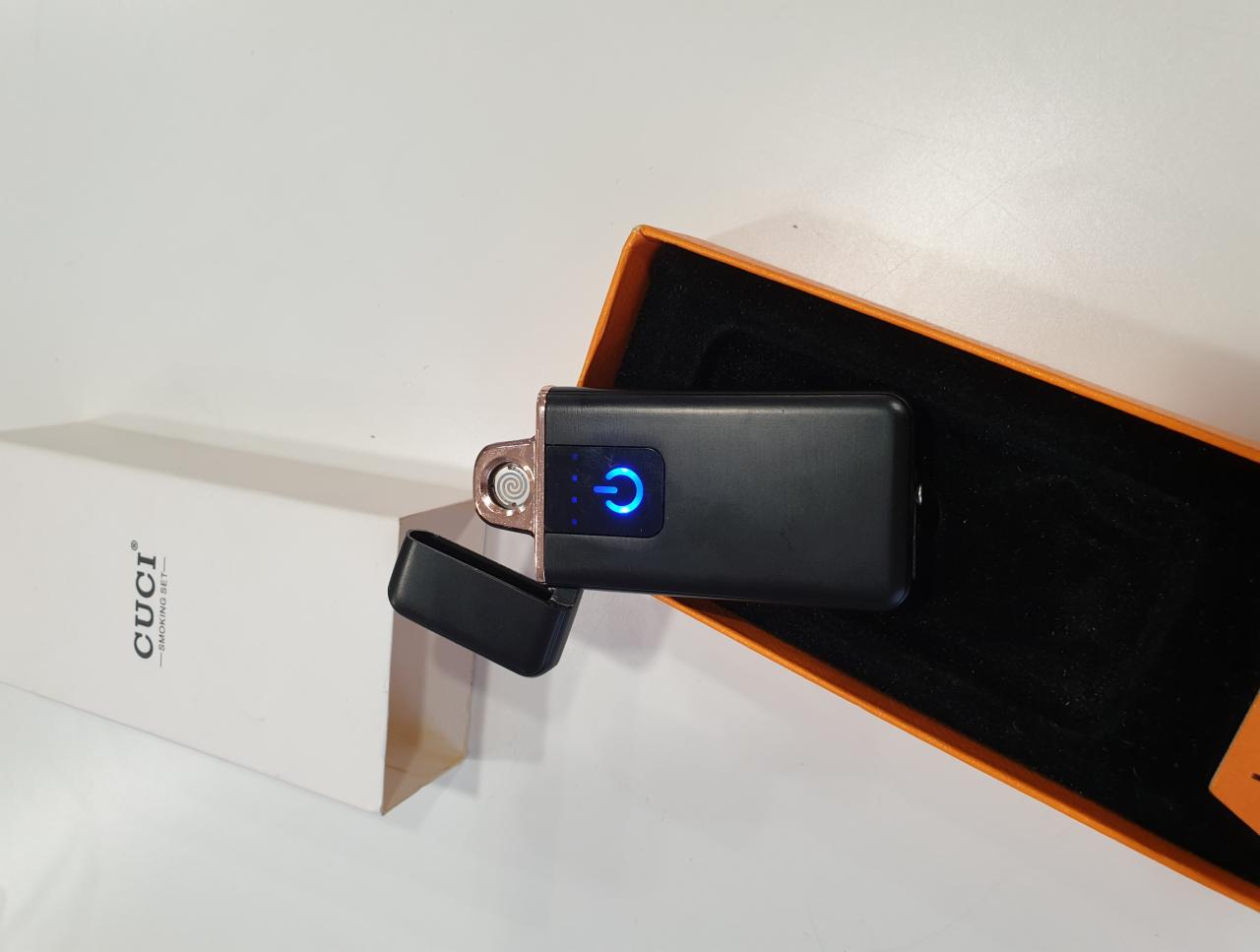 Elegancki zestaw upominkowy: czarna zapalniczka plazmowa z niebieskim podświetleniem w pomarańczowym pudełku prezentowym, obok pudełko marki CUCI.