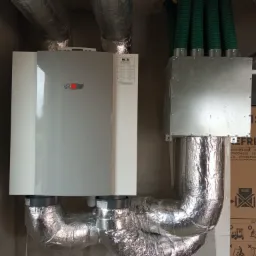 Zamontowana centrala wentylacyjna Wolf z przewodami wentylacyjnymi w izolacji aluminiowej i zielonymi rurami wentylacyjnymi, widoczna kartonowa skrzynia obok.